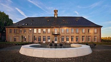 Historisches Gebäude mit einem Uhrturm, umgeben von einem runden Sitzbereich und einer gepflasterten Fläche.