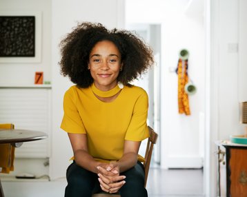 Eine lächelnde Frau mit lockigem Haar sitzt in einem hellen Raum. Sie trägt ein gelbes Oberteil und hat die Hände auf den Knien gefaltet.