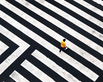 Eine Person in gelber Kleidung geht über ein geometrisches Zebrastreifenmuster aus schwarzen und weißen Linien.